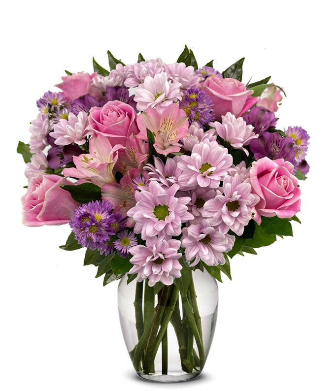 A full bouquet of pink roses, lavender daisy poms, pink alstroemeria, and purple asters in a clear glass vase with leafy greenery.