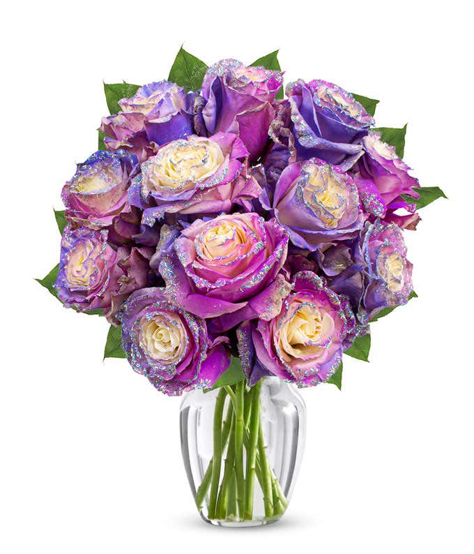 A bouquet of pink, purple, and cream roses with glittered petal edges arranged in a clear glass vase with green leaves.