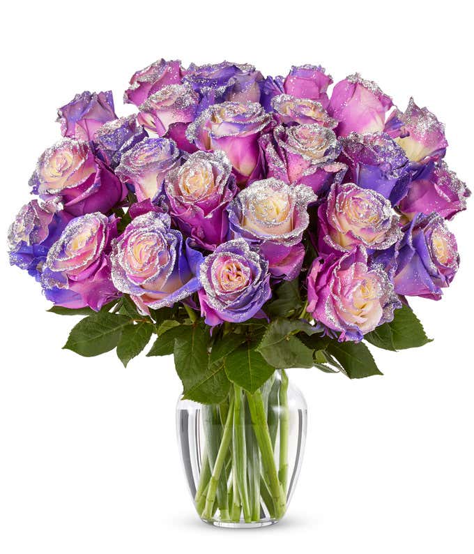 Two dozen purple roses with iridescent glitter on the petals, arranged with fresh green leaves in a clear glass vase.