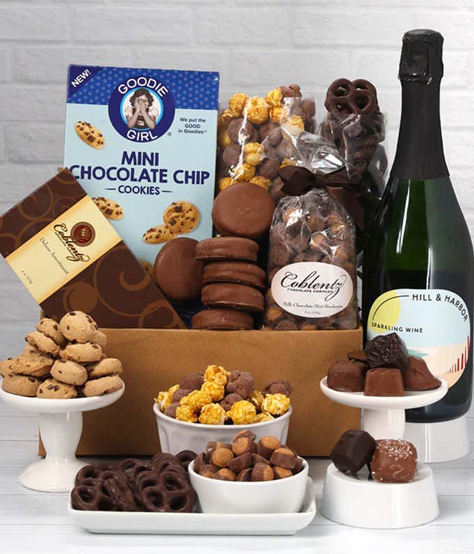 a gift box and a bottle of sparkling wine on a kitchen counter with a glass of wine and some chocolate treats on plates. Gift box includes cookies, chocolates, peanut butter chocolates, chocolate oreos, and chocolate pretzels