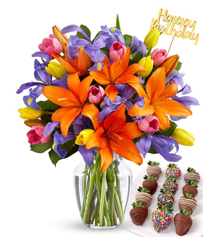 Bright bouquet of orange lilies, purple irises, and multicolored tulips in a clear glass vase with a gold Happy Birthday pick, paired with a plate of chocolate-covered strawberries featuring swizzled and rainbow sprinkle designs.