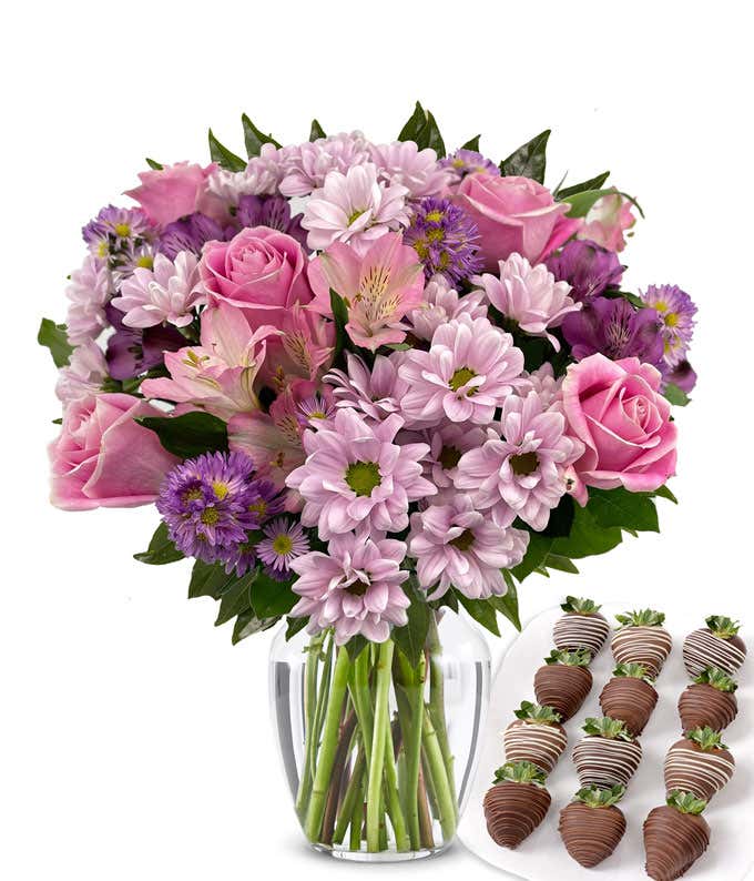 Elegant bouquet of pink roses, lavender cushion poms, purple alstroemeria, pink alstroemeria, and purple asters in a clear glass vase, paired with swizzled chocolate-covered strawberries.