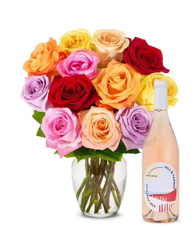 A bouquet of roses in various colors including red, pink, orange, yellow, and purple, neatly arranged in a clear glass vase. Beside it is a slender bottle of rose wine with a pale pink label from California, 2023, showcasing an elegant design.