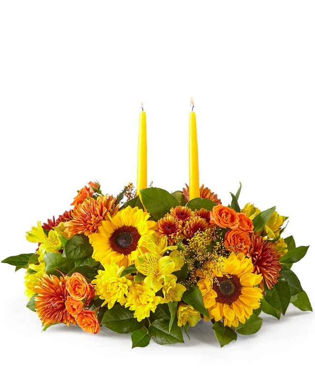 A cheerful floral arrangement featuring sunflowers, butterscotch mums, yellow alstroemeria, butterscotch cushion poms, orange spray roses, yellow cushion poms, red-tinted solidago, and seasonal greens, complemented by yellow taper candles.