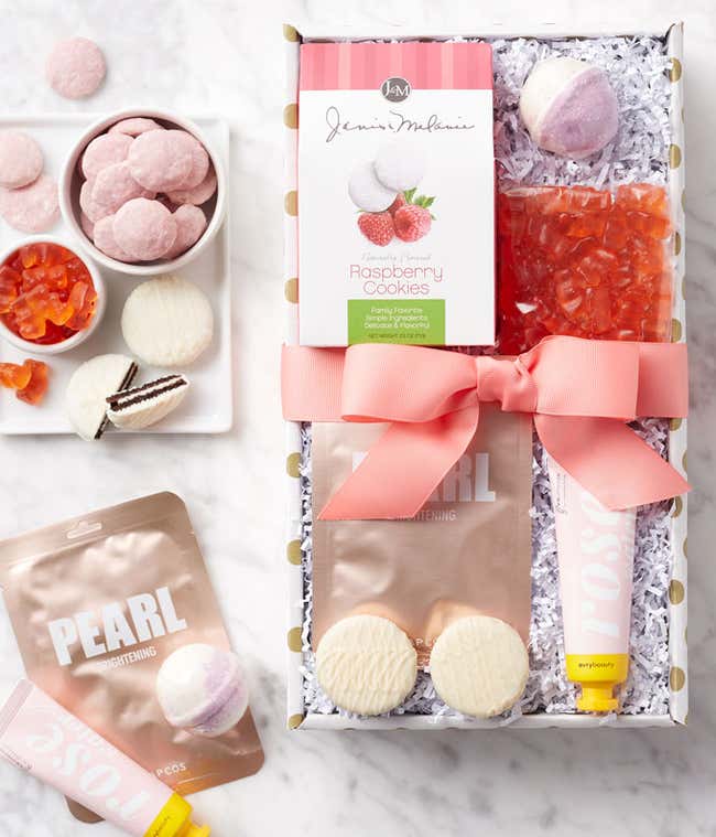 Gift box featuring Champagne Gummies, Raspberry Cookies, White Chocolate Sandwich Cookies, Rose Hand Cream, a Face Mask, and a Bath Bomb, all arranged with a pink ribbon bow.