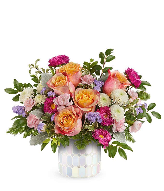 Elegant floral arrangement featuring peach and orange roses, pink spray roses, hot pink Matsumoto asters, white poms, lavender statice, and assorted greenery in a pastel hexagon-patterned ceramic container.