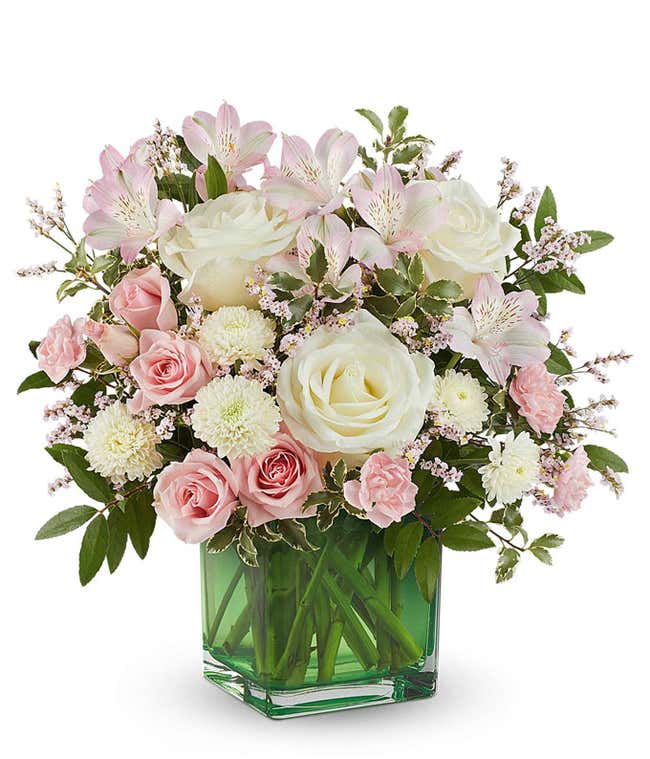 Soft pastel floral arrangement featuring cr&egrave;me roses, pink spray roses, pink alstroemeria, miniature pink carnations, white button spray mums, delicate filler flowers, and lush greenery in a green glass cube vase.
