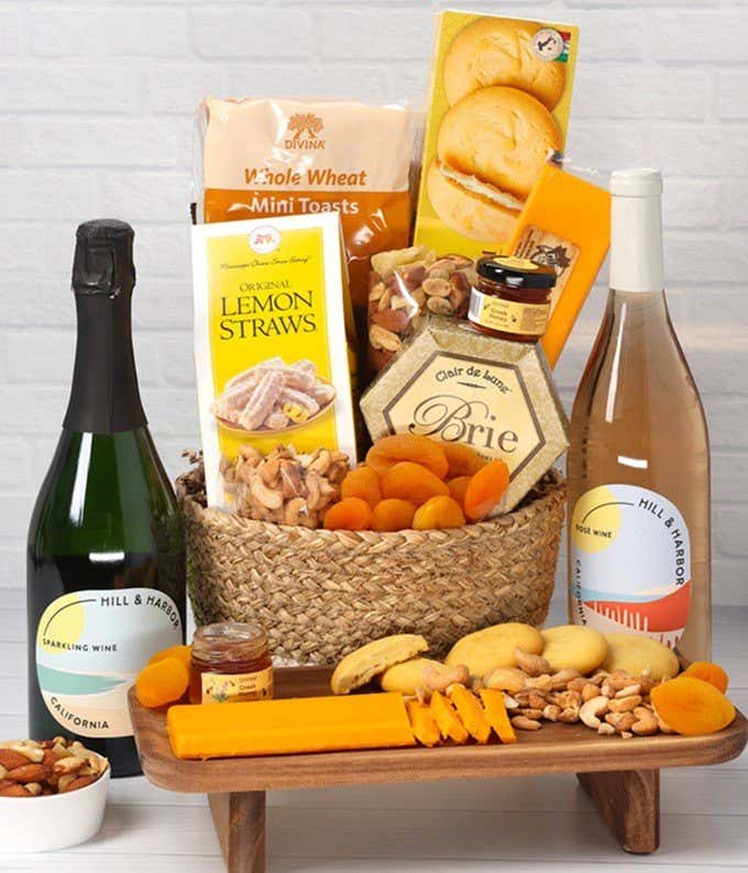 A two-toned woven basket filled with two cheeses, lemon flavored baked goods, crackers, dried apricots, gourmet nuts, and honey, paired with two bottles of wine - Sparkling & Rosé.